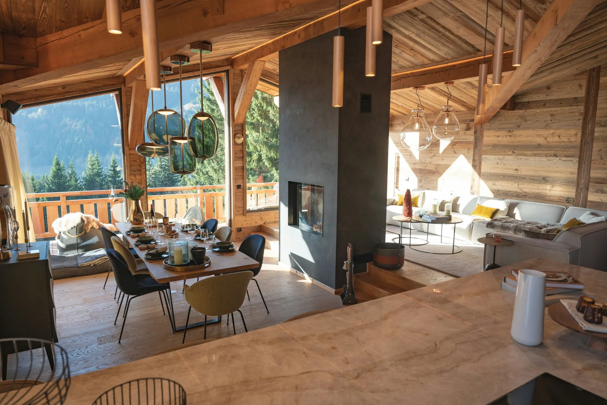 Pièce de vie d'un chalet en vieux bois, cheminée centrale et grandes baies ouvertes sur la forêt